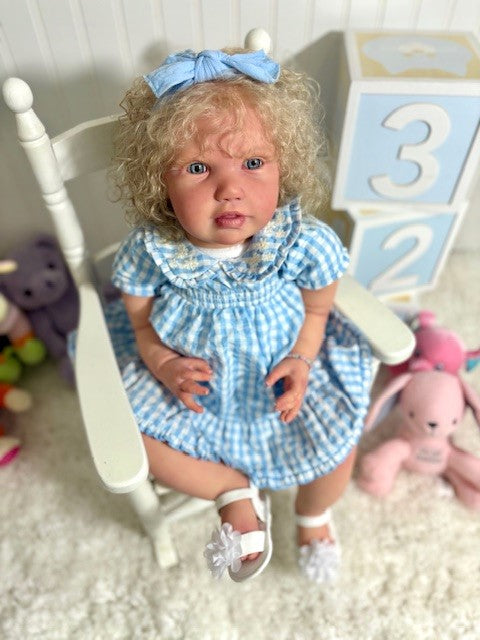 Reborn Baby doll in a blue checkered dress sitting on a white chair with toys around.